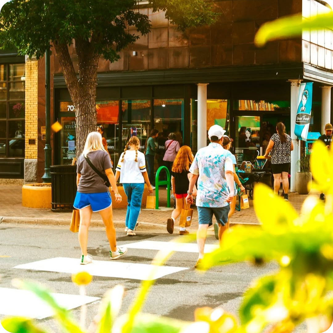 A group of four people crosses a street in a busy area, with a storefront in the background and a green bike rack nearby.